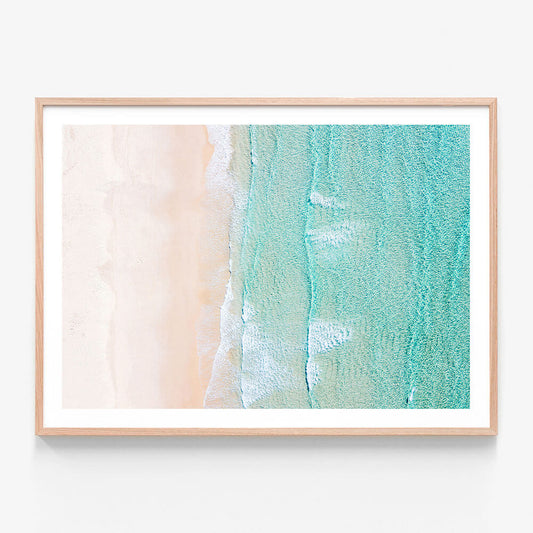 A framed coastal wall art print featuring an overhead photo of the beach with turquoise water and clean sand