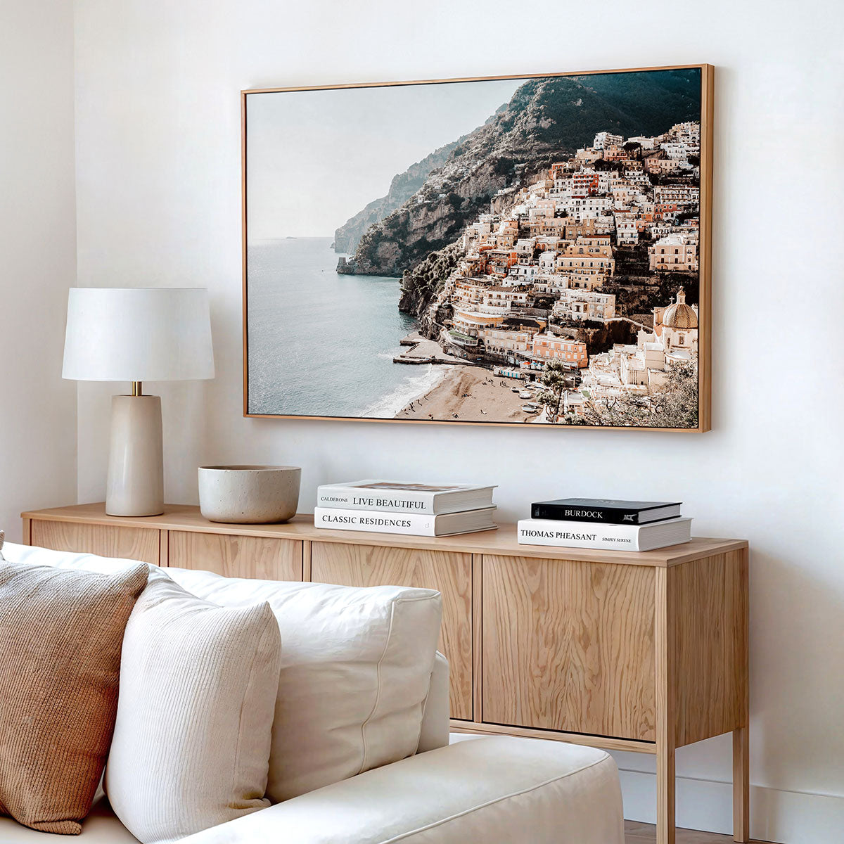 Living room with a framed canvas artwork of a coastal town scene from Europe. With a wooden sideboard and white sofa,.