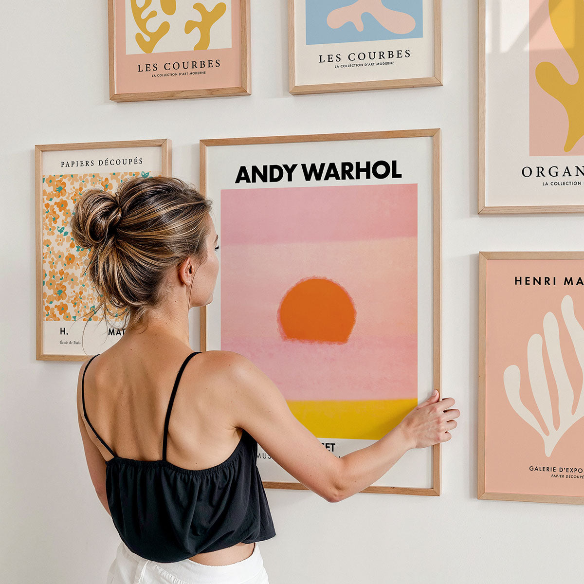 Woman arranging framed posters on a gallery wall with 'Andy Warhol' and other abstract art pieces by famous artists.