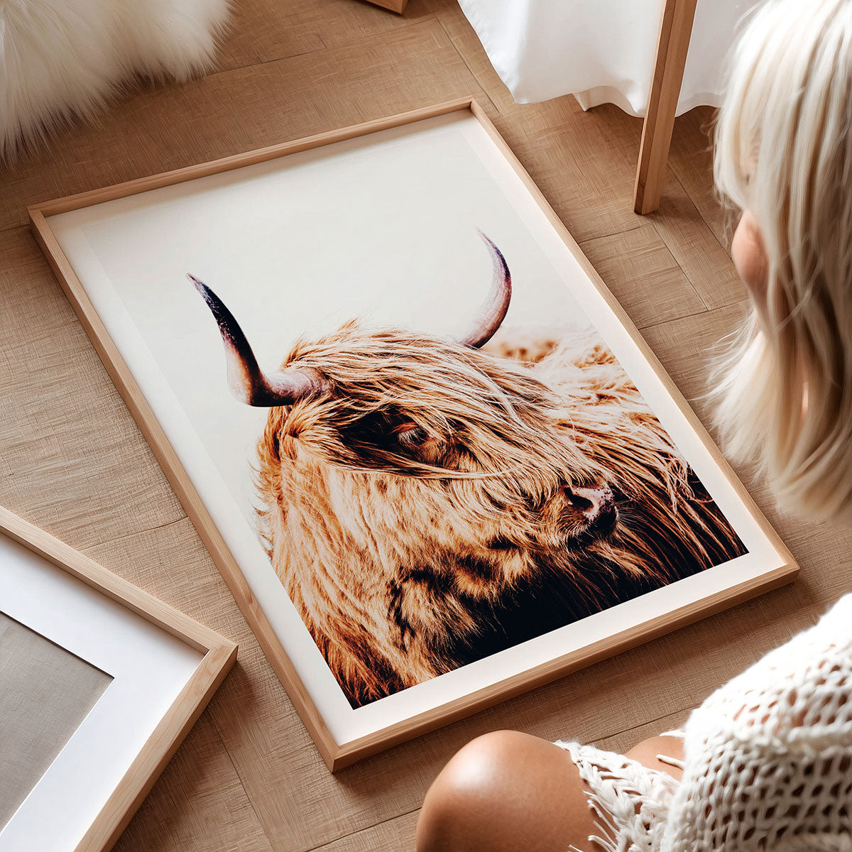 Framed artwork print of a highland cow on a wooden floor with a person sitting nearby.