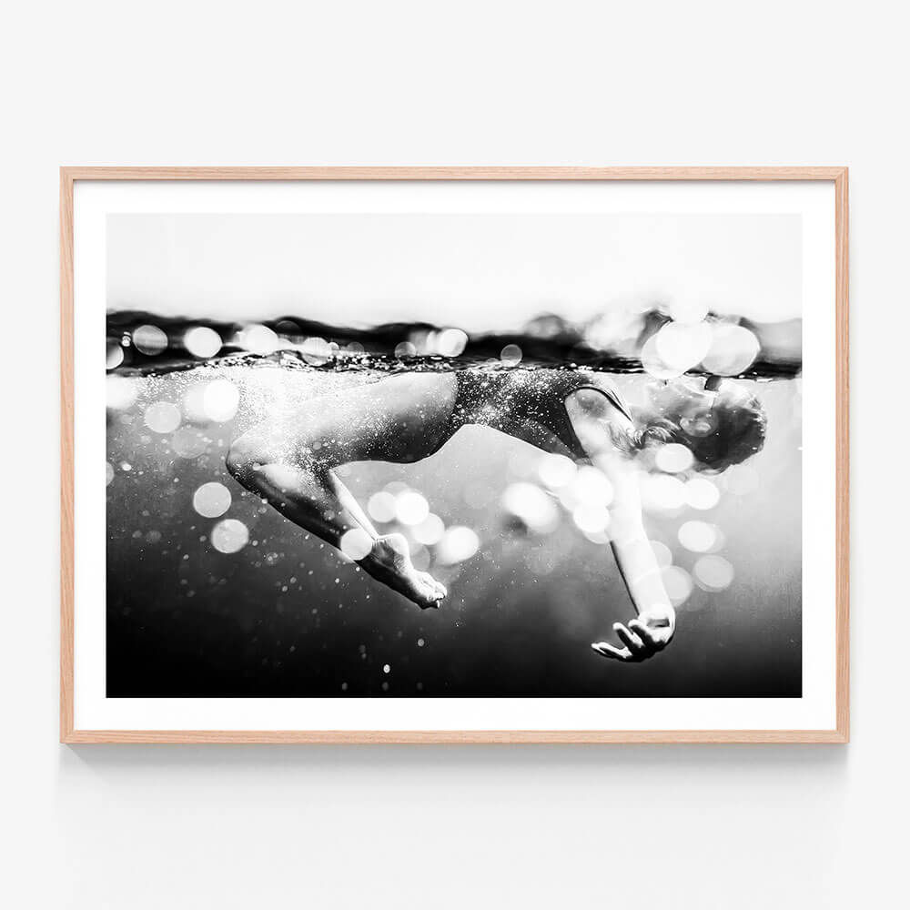 This image titled "Immerse" features a striking black and white photograph of a person swimming underwater. The framed print captures the essence of motion and breath, with bubbles rising to the surface indicating the action of breathing and the playful nature of being in water.
