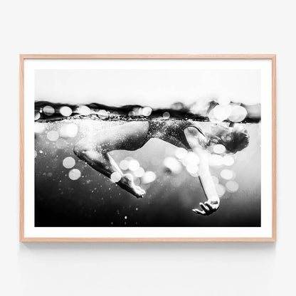 This image titled "Immerse" features a striking black and white photograph of a person swimming underwater. The framed print captures the essence of motion and breath, with bubbles rising to the surface indicating the action of breathing and the playful nature of being in water.