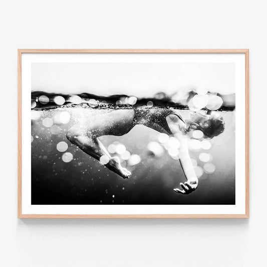 This image titled "Immerse" features a striking black and white photograph of a person swimming underwater. The framed print captures the essence of motion and breath, with bubbles rising to the surface indicating the action of breathing and the playful nature of being in water.