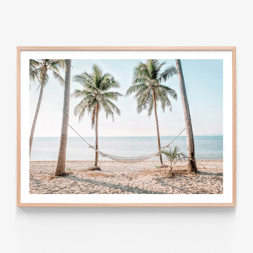A framed photo print featuring a hammock strung between two palm trees on a beach, with the ocean in the background.