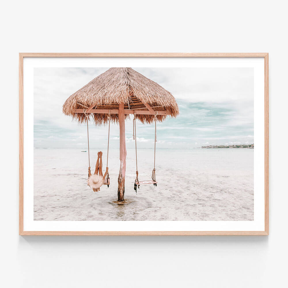 A coastal framed print featuring a swing hanging from a straw umbrella on a beach, with the ocean in the background.