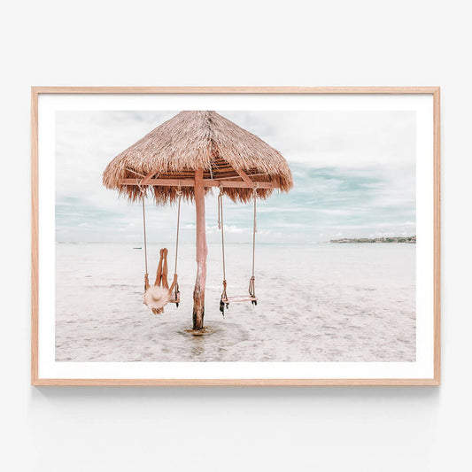 A coastal framed print featuring a swing hanging from a straw umbrella on a beach, with the ocean in the background.