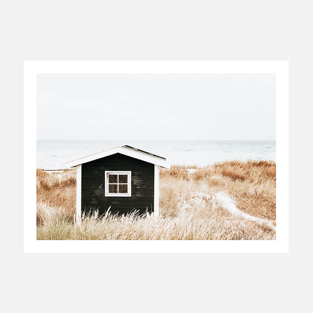A wall art print of a small house on a coastal beach with tall grasses in the foreground, predominantly in black, white, and beige colors.