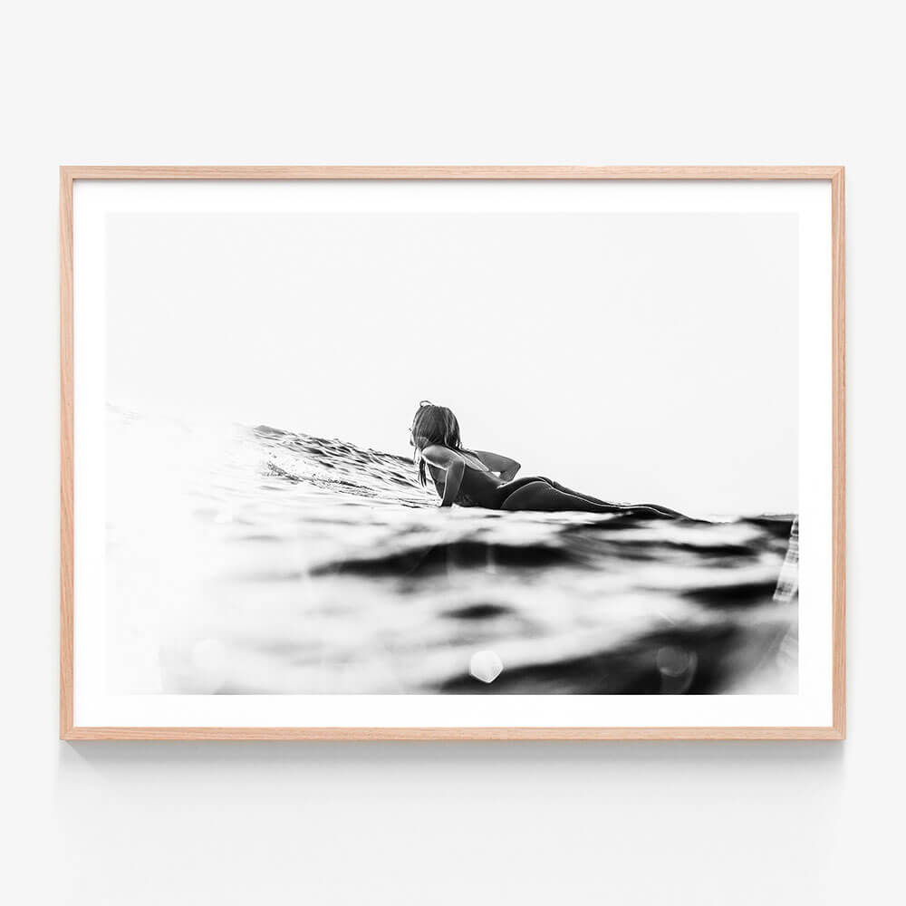 A framed art print featuring a black and white image of a person lying on a surfboard, paddling in the ocean.