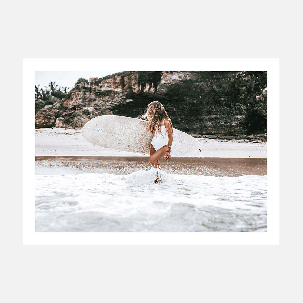 A photo wall art print featuring a woman carrying a surfboard on their head while walking along a beach with ocean waves and rocks in the background.