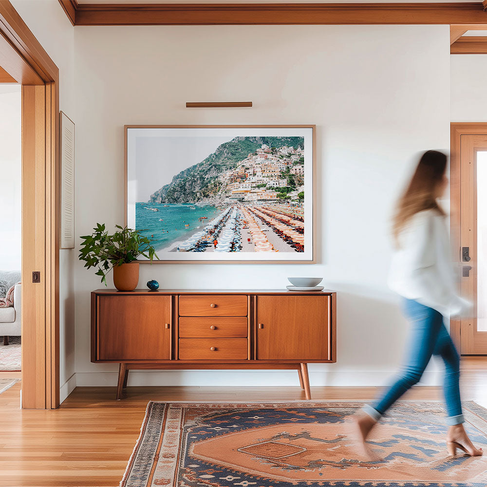 Living room with a large framed picture of an Italian coastal scene of the beach at Positano. A wooden sideboard, and a person walking by.