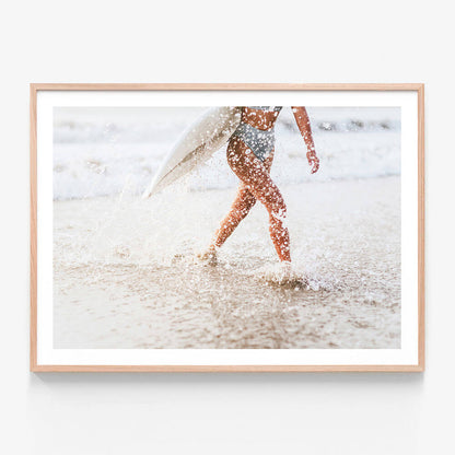 A framed photo wall art print featuring a woman carrying a surfboard while walking on a sandy beach with water splashing around.