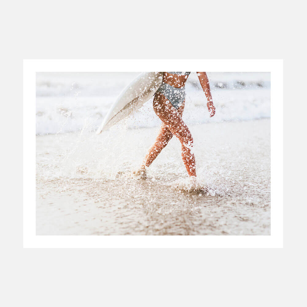 A photo print person carrying a surfboard while walking on a sandy beach with water splashing around.