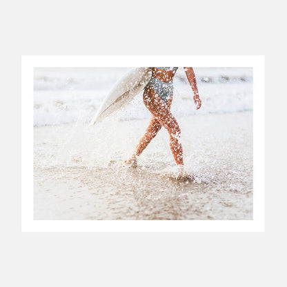 A photo print person carrying a surfboard while walking on a sandy beach with water splashing around.