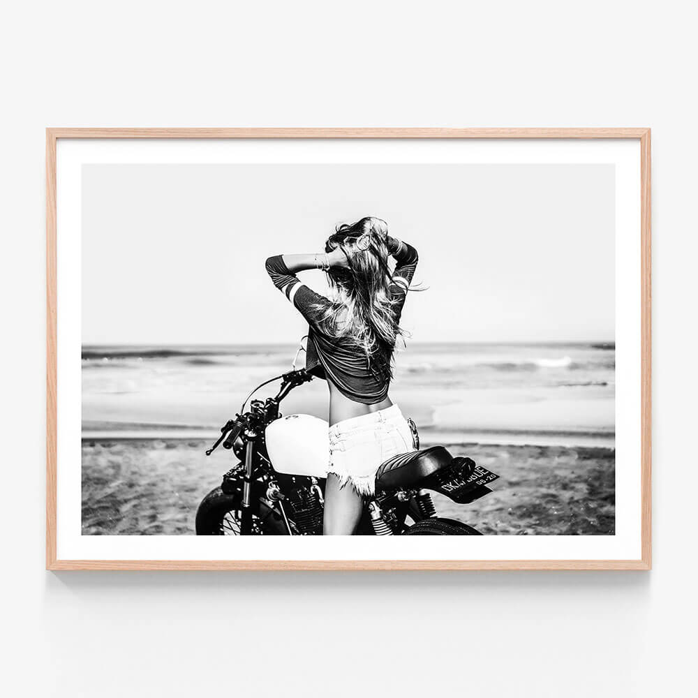 This framed art print features a striking black and white photograph capturing a vivid beach scene. A woman is seen sitting on a motorcycle with a backdrop of the ocean and sand dunes.