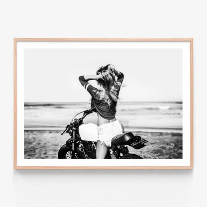 This framed art print features a striking black and white photograph capturing a vivid beach scene. A woman is seen sitting on a motorcycle with a backdrop of the ocean and sand dunes.