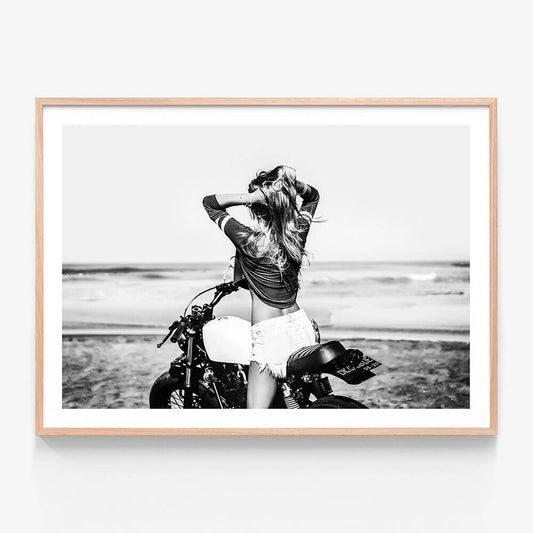 This framed art print features a striking black and white photograph capturing a vivid beach scene. A woman is seen sitting on a motorcycle with a backdrop of the ocean and sand dunes.