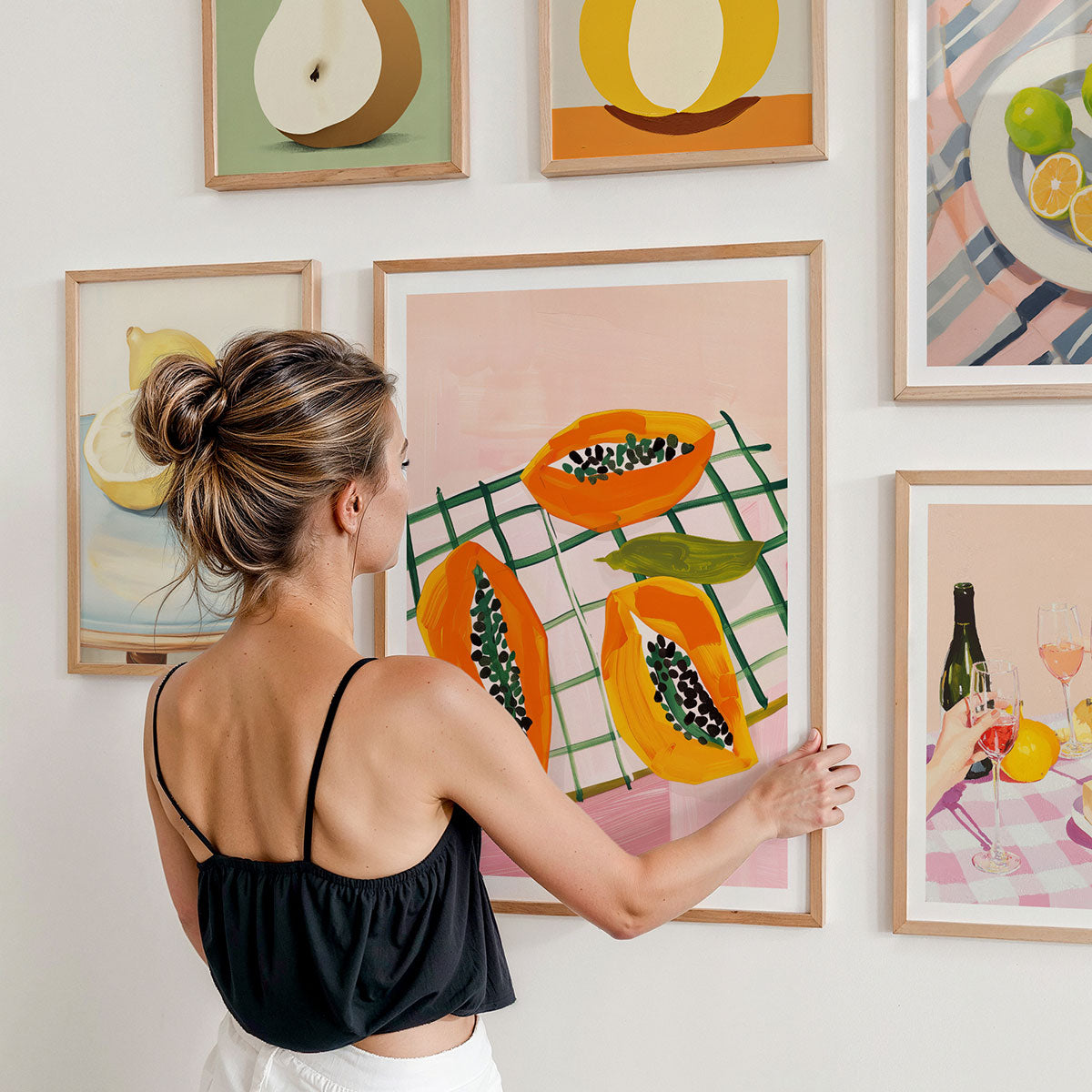 A woman arranging framed artworks on a gallery wall featuring colourful still life illustrations.