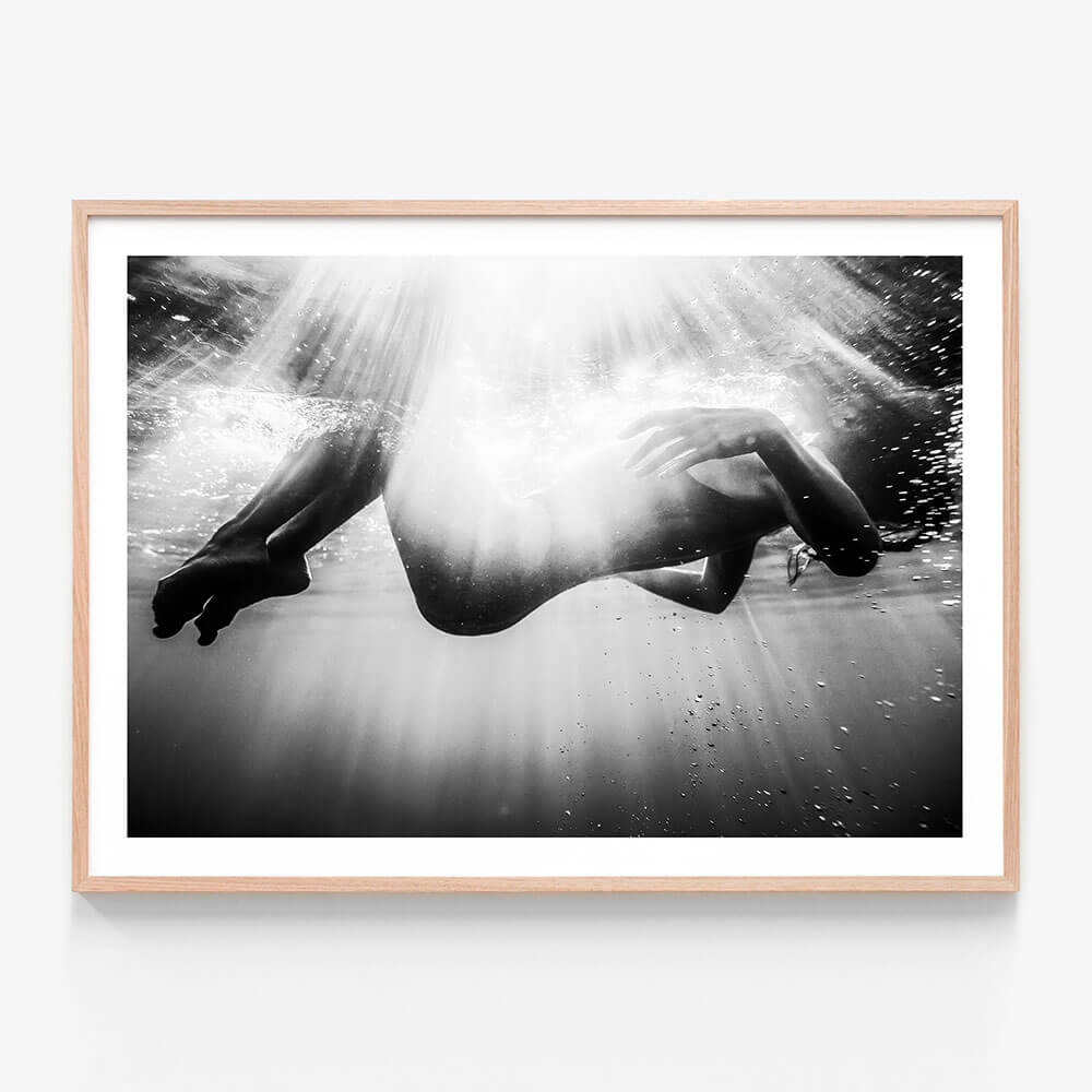 This framed art print features a striking black and white photograph titled "Submerge." It captures the ethereal and powerful moment of a person underwater, reaching towards the surface.