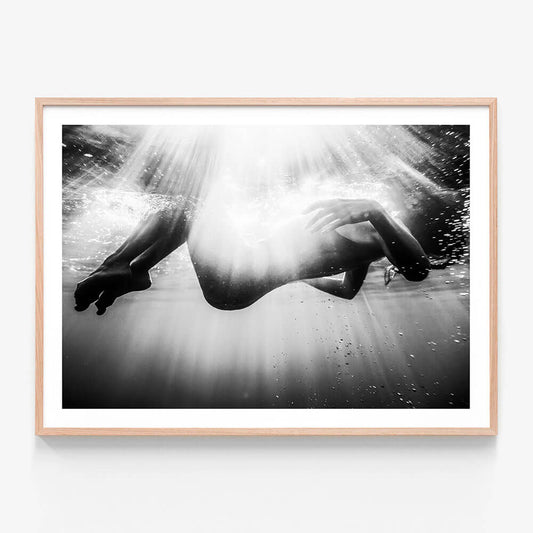 This framed art print features a striking black and white photograph titled "Submerge." It captures the ethereal and powerful moment of a person underwater, reaching towards the surface.