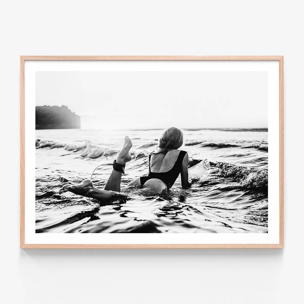 A framed black and white artwork of a person lying on a surfboard in the ocean, with waves around and a sunset in the background.