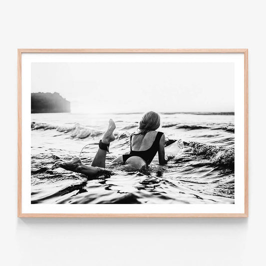 A framed black and white artwork of a person lying on a surfboard in the ocean, with waves around and a sunset in the background.
