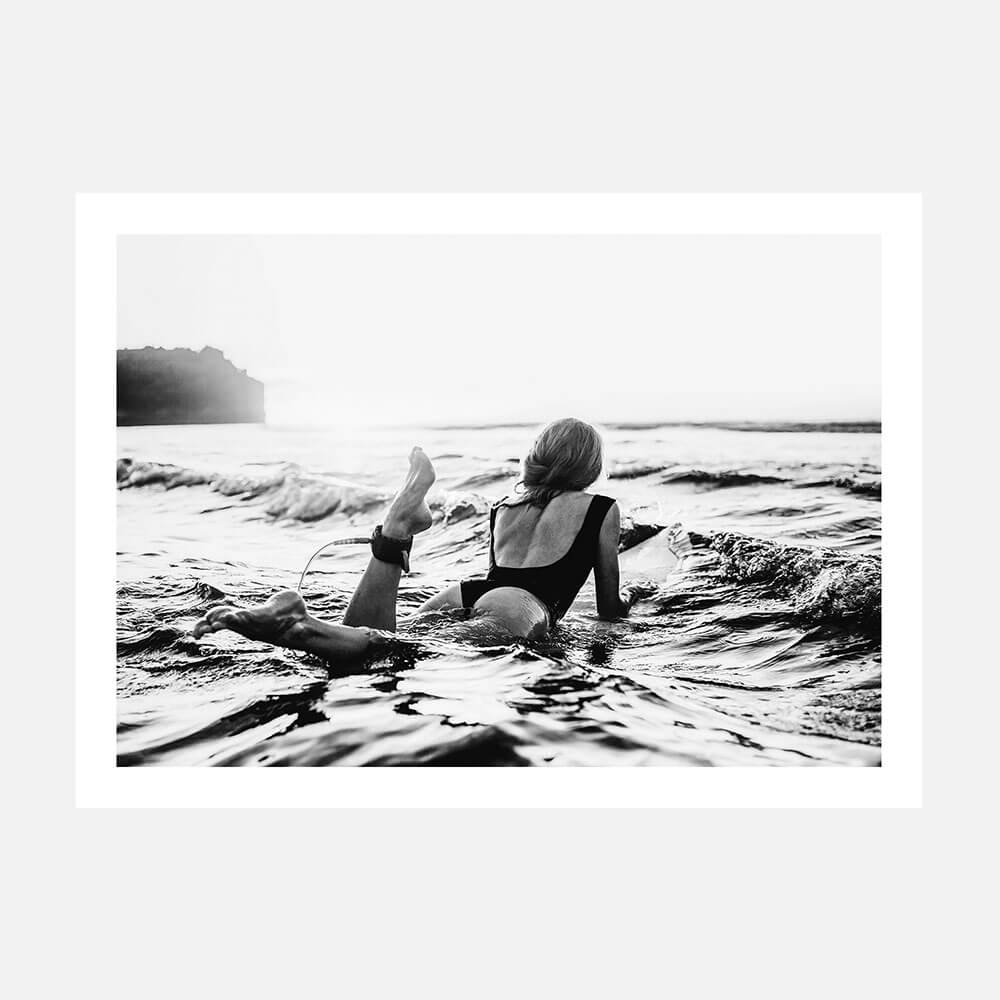A black and white photo art print of a person lying on a surfboard in the ocean, with waves around and a sunset in the background.