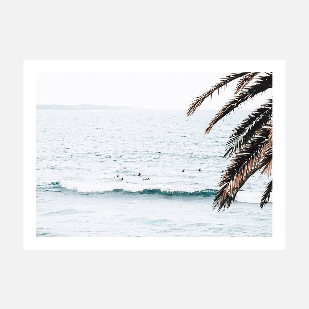 A print featuring a serene beach scene with surfers in the ocean, palm trees in the foreground, and a predominantly white and blue color palette.