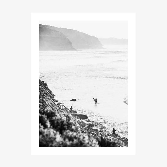 A black and white photograph art print of surfers in the ocean at Bells beach with a coastal landscape in the background.