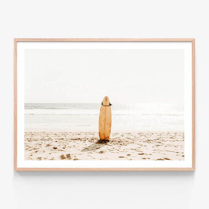 A framed photo wall art print featuring a surfboard standing upright on a sandy beach with the ocean in the background.