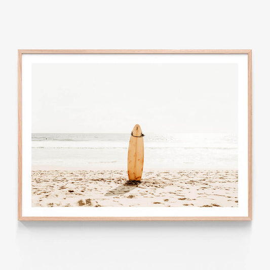 A framed photo wall art print featuring a surfboard standing upright on a sandy beach with the ocean in the background.