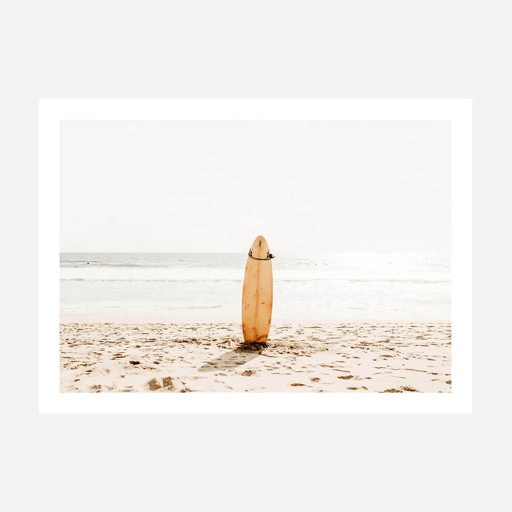 A coastal style photo wall art print featuring a surfboard standing upright on a sandy beach with the ocean in the background.