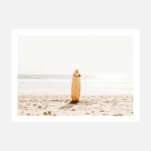 A coastal style photo wall art print featuring a surfboard standing upright on a sandy beach with the ocean in the background.