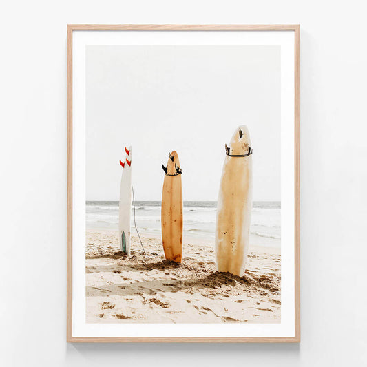 This image features a charming artwork titled "Three Boards," perfect for adding a coastal vibe to any space. The artwork displays three surfboards standing upright on a sandy beach, each with distinct and playful decorations.