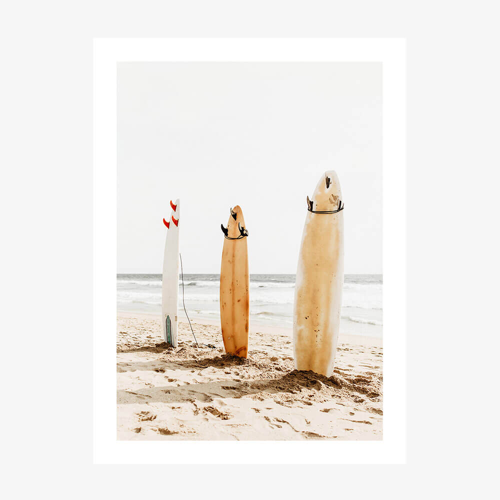 This image features a charming artwork titled "Three Boards," perfect for adding a coastal vibe to any space. The artwork displays three surfboards standing upright on a sandy beach, each with distinct and playful decorations.