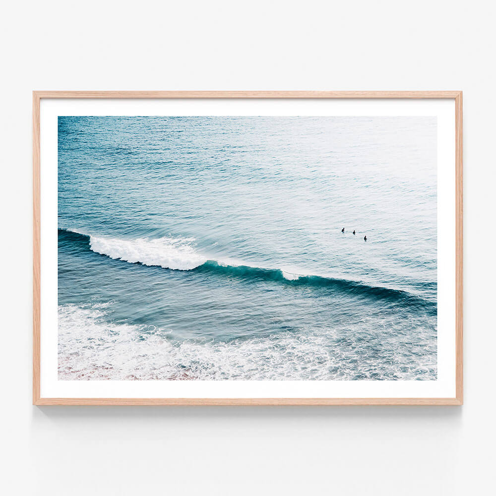 A framed photo print featuring a coastal scene with gentle waves and three surfers in the distance.