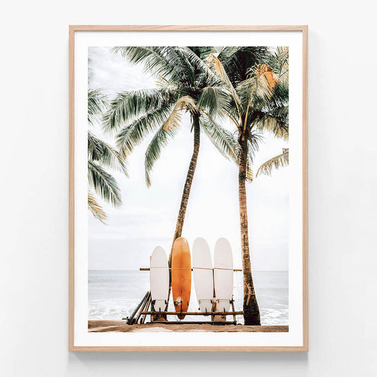 A framed wall art print featuring a tropical beach scene with palm trees, a bench, and multiple surfboards placed against a white backdrop.