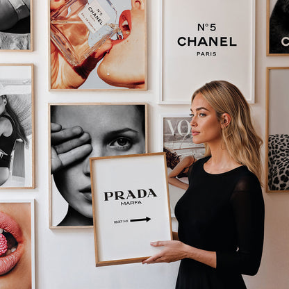Woman holding a typographic framed print with the text 'Prada' against a wall of framed art and typography posters.