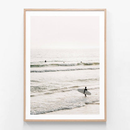 A framed photo wall art print featuring a coastal scene with a person holding a surfboard, standing in the shallow ocean waters with waves and a clear sky in the background.