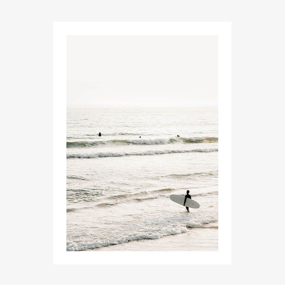 A photo print of a coastal scene with a person holding a surfboard, standing in the shallow ocean waters with waves and a clear sky in the background.
