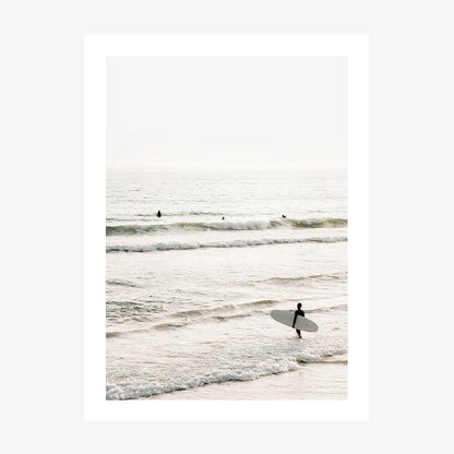A photo print of a coastal scene with a person holding a surfboard, standing in the shallow ocean waters with waves and a clear sky in the background.