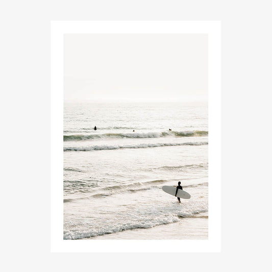 A photo print of a coastal scene with a person holding a surfboard, standing in the shallow ocean waters with waves and a clear sky in the background.