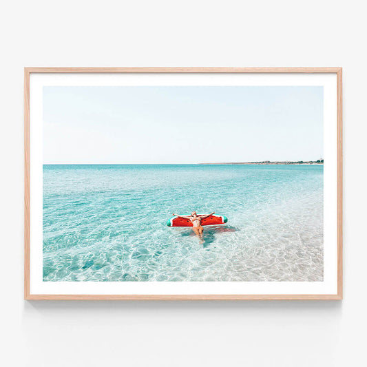 A framed photo wall art print featuring a  person floating on a red floatie in a clear blue ocean.