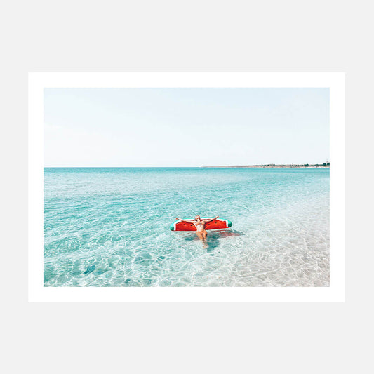 A photo wall art print featuring a woman floating on a watermelon floatie in a clear blue ocean.