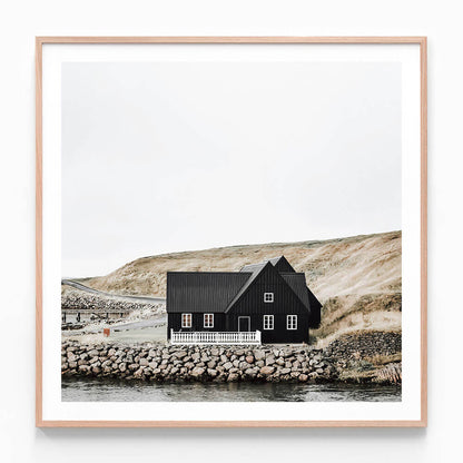 A framed photography print of a black wooden house with a thatched roof situated by a body of water, with a rocky shore in the foreground.