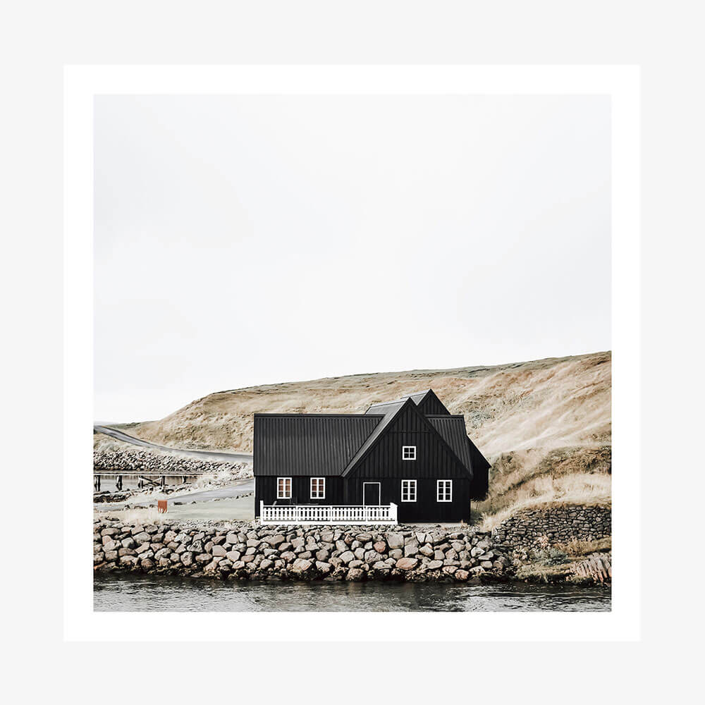 A photo art print of a black wooden house with a thatched roof situated by a body of water, with a rocky shore in the foreground.