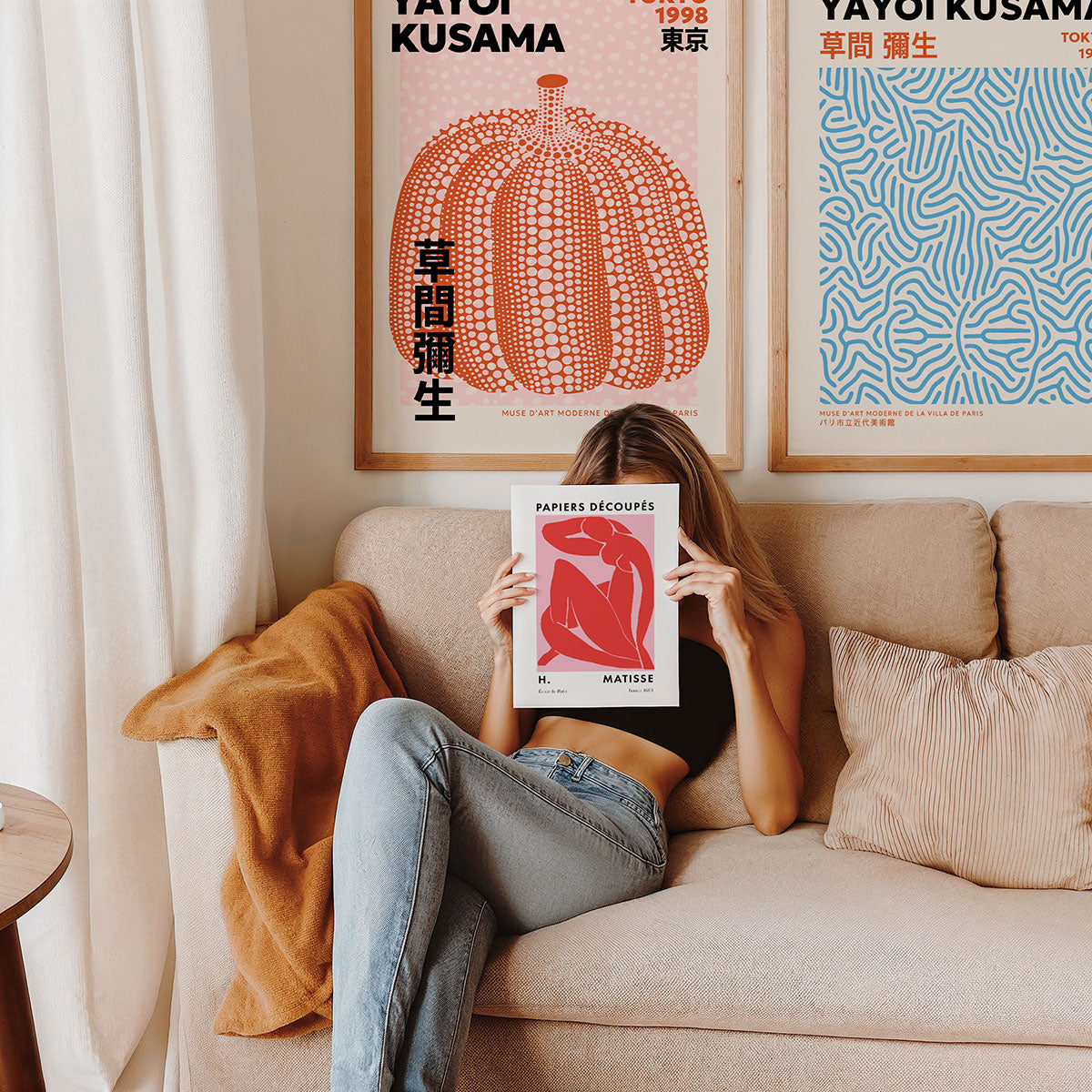 Person sitting on a couch holding a Matisse poster in front of their face, with tow Yayoi Kusama framed prints on the wall.